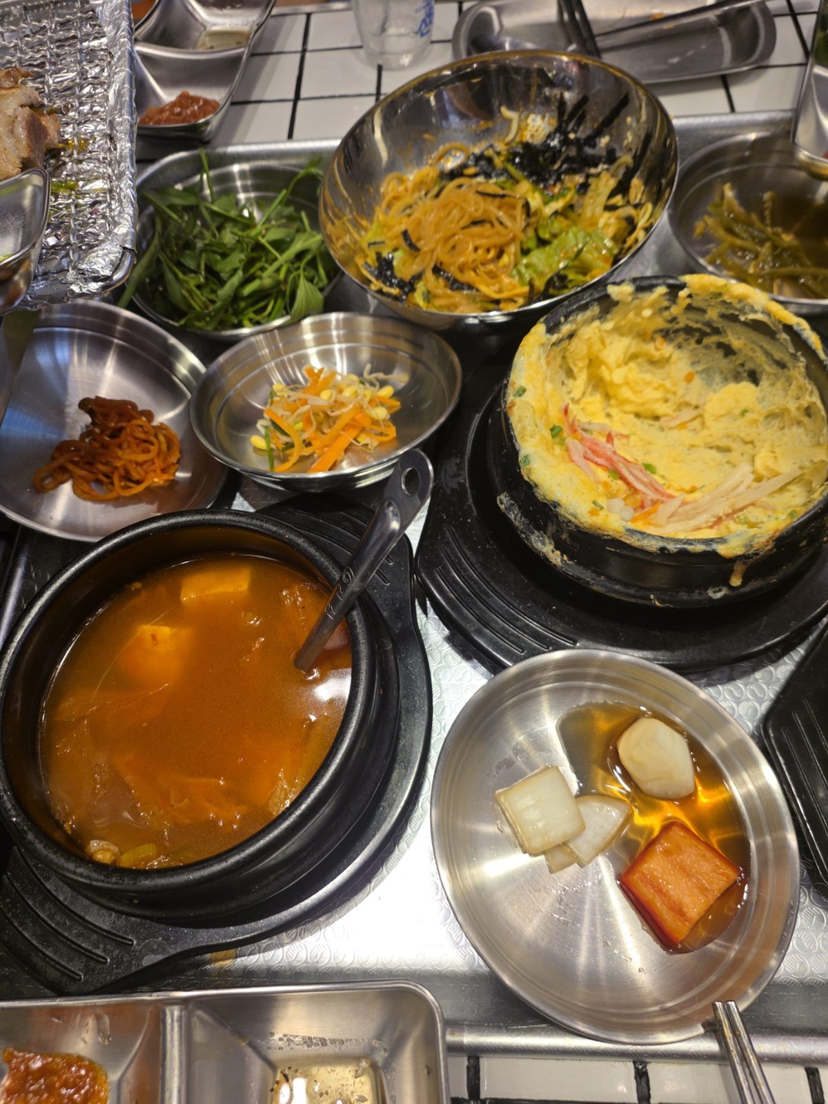 Bàn món ăn kèm đang dùng dở gồm rau cần nước, mì trộn, rau mầm, đồ chua, canh kimchi và trứng hấp có thanh cua